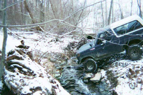 Dark blue Ford Bronco with a white top navigating a snowy stream, angled from the front-right