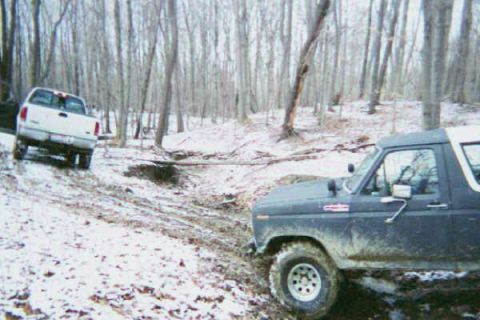 1986 Ford Bronco - Two 4x4 vehicles navigate a challenging off-road course in a wooded area during winter.