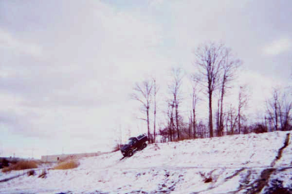 Dark pickup truck climbing a snow-covered hill with bare trees in the background