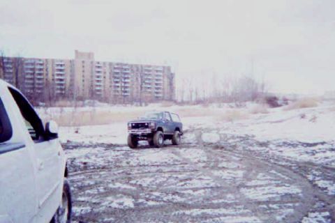 1986 Ford Bronco - A dark blue SUV navigates a snowy and muddy terrain with a large apartment building in the background.