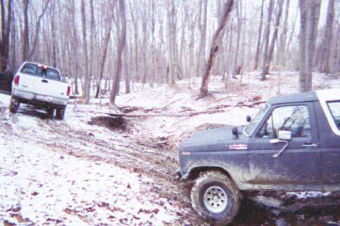 1986 Ford Bronco - Two trucks, a Ford Bronco and an F-150, navigate a muddy, snowy trail in a wooded area.