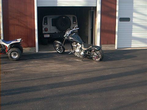 1986 Ford Bronco - A custom chopper motorcycle and an ATV are parked outside a garage, with a white SUV visible inside.