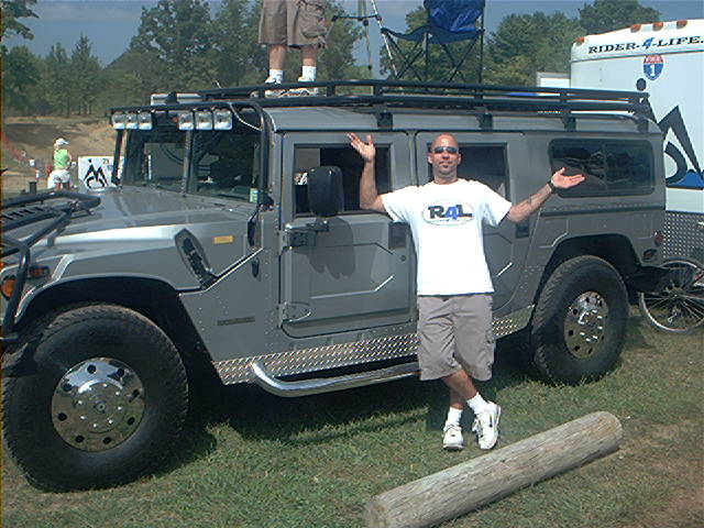 Man poses beside silver Hummer H1 with roof rack and light bar on grass