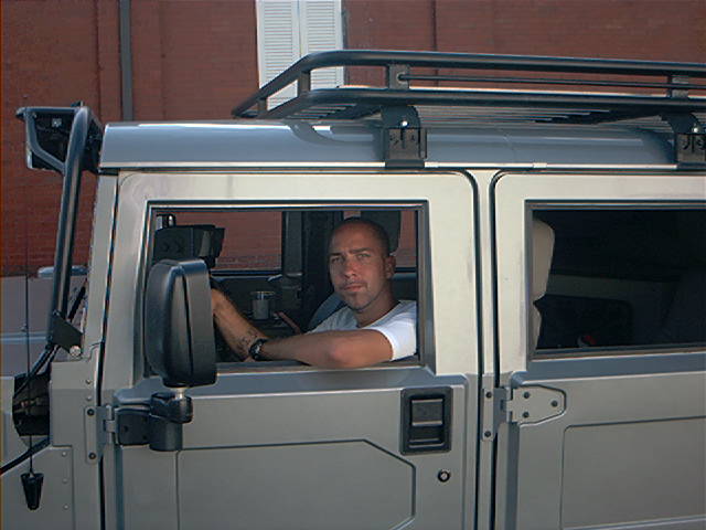 Silver Hummer-style SUV with roof rack, driver leaning arm out the window, brick building behind