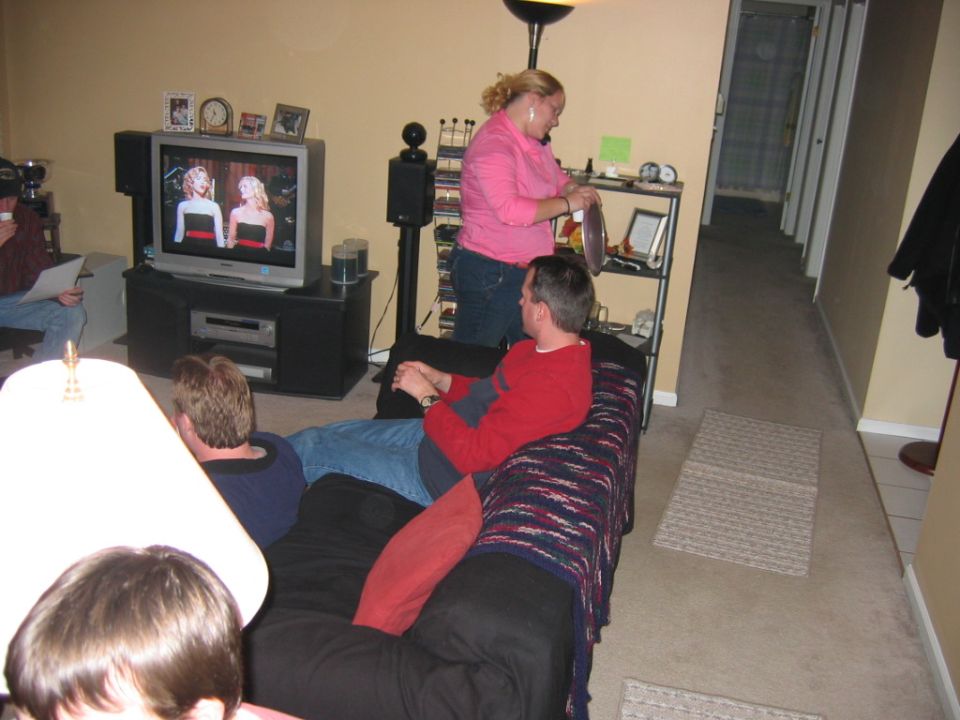People in a living room watching TV. A woman in pink stands near a shelf