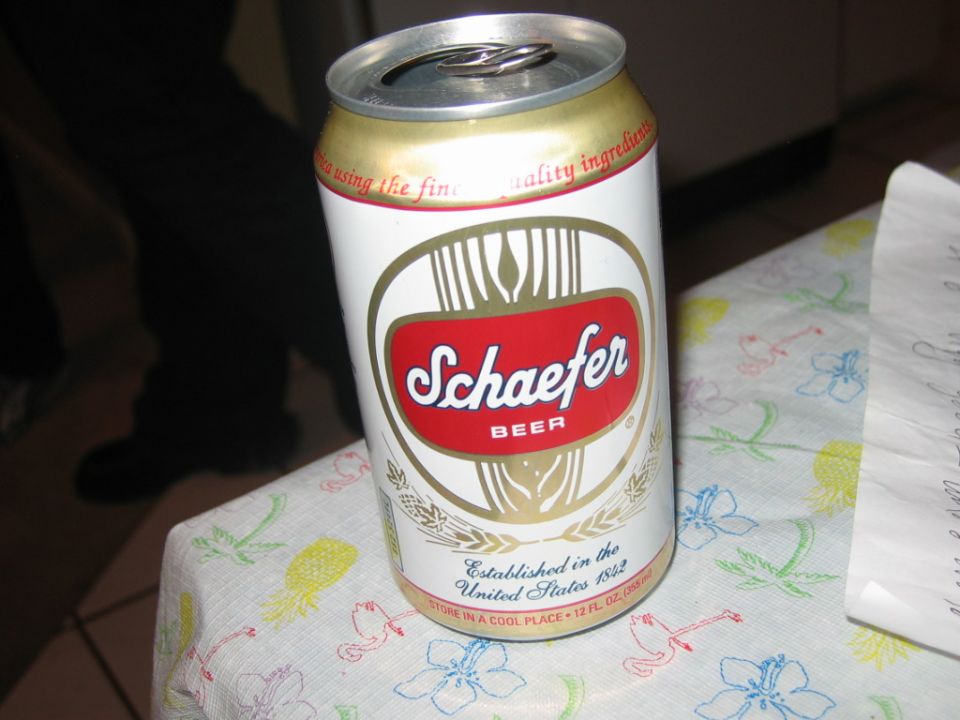 Schaefer beer can on a table with a floral tablecloth. Legs of a person are visible in the background