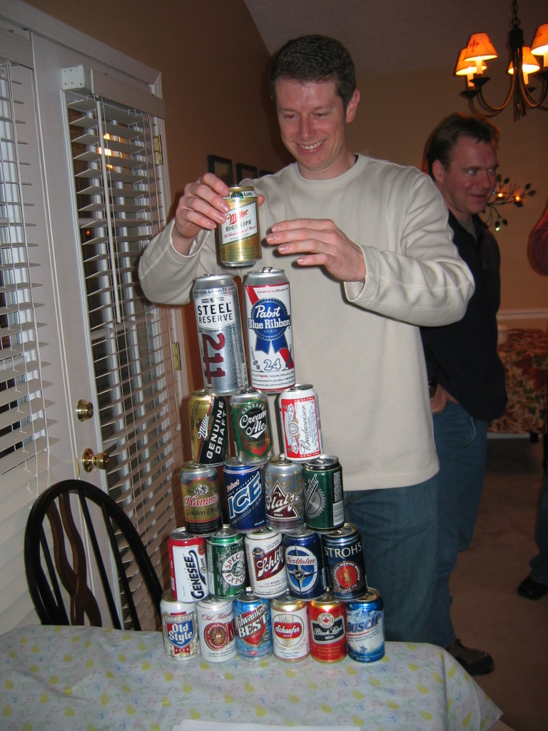 Man stacking beer cans into a pyramid, with another man in the background