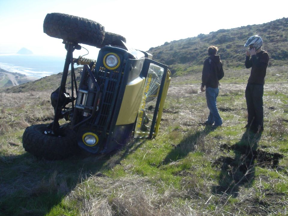 Yellow off-road rig with roll cage tipped onto its side on a grassy hill; two people stand nearby