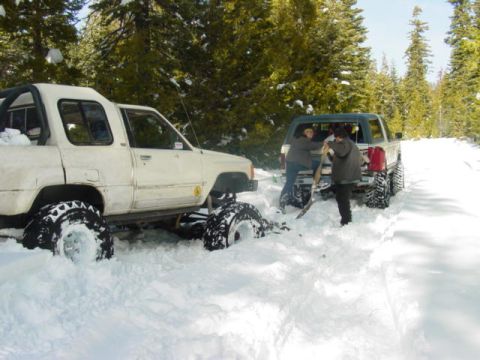 1986 Ford Bronco - Two trucks are stuck in deep snow in a wooded area, with people attempting to dig one out.