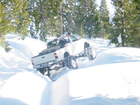 1986 Ford Bronco - A white pickup truck navigates a snowy terrain with its large tires, while a person stands in the truck bed.