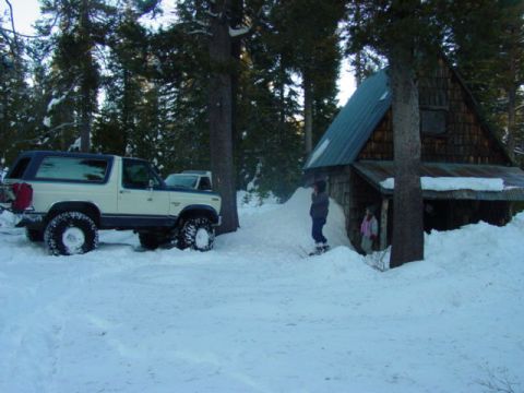 1986 Ford Bronco - A 1986 Ford Bronco is parked in a snowy landscape next to a rustic cabin, with two people visible near the entrance.