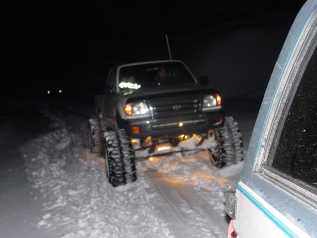 Truck with tracks driving in snow at night, another vehicle to the right