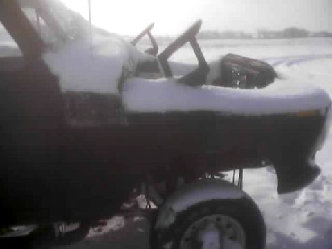 1979 Ford Bronco - A dark vehicle sits partially covered in snow in a field, with only a portion of the front and a wheel visible.