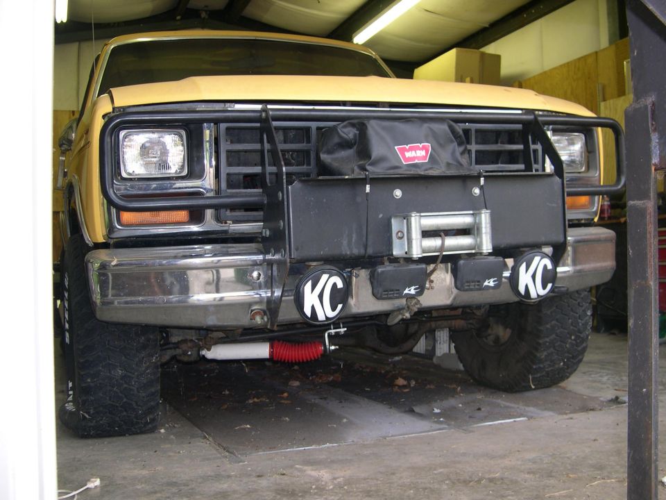 Yellow 1980s Ford Bronco front view with winch, bull bar, and KC lights. Supermotors.net logo visible