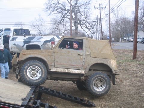 1987 Suzuki Samurai - A driver holds a beer while operating a muddy 1987 Suzuki Samurai, which is partially on a trailer, with another truck visible behind it.