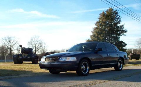 1998 Ford Crown Victoria - A dark blue sedan is parked on a road with a tank in the background under a clear sky.