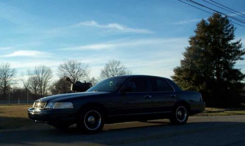 1998 Ford Crown Victoria - A dark 1998 Ford Crown Victoria is parked outdoors under a bright blue sky with scattered clouds.