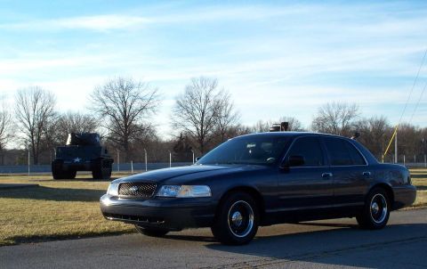 1998 Ford Crown Victoria - A 1998 Ford Crown Victoria is parked next to a military tank under a partly cloudy sky.