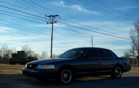 1998 Ford Crown Victoria - A dark sedan is parked near a utility pole, with military vehicles in the distance under a blue sky.