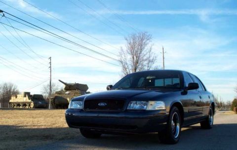 1998 Ford Crown Victoria - A dark sedan is parked on a road, with two military tanks visible in the background under a blue sky.