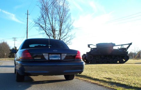 1998 Ford Crown Victoria - A dark 1998 Ford Crown Victoria is parked on a road next to a military tank under a bright blue sky.