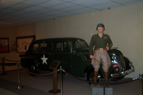 1998 Ford Crown Victoria - A vintage dark green sedan is displayed next to a mannequin dressed as a soldier in a museum setting.