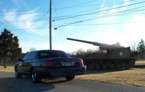 1998 Ford Crown Victoria - A dark sedan is parked on a road with a military self-propelled howitzer in the background under a cloudy sky.