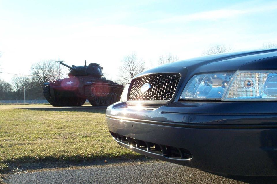 Close-up of dark blue Ford Crown Victoria front grille and headlight, tank on display behind