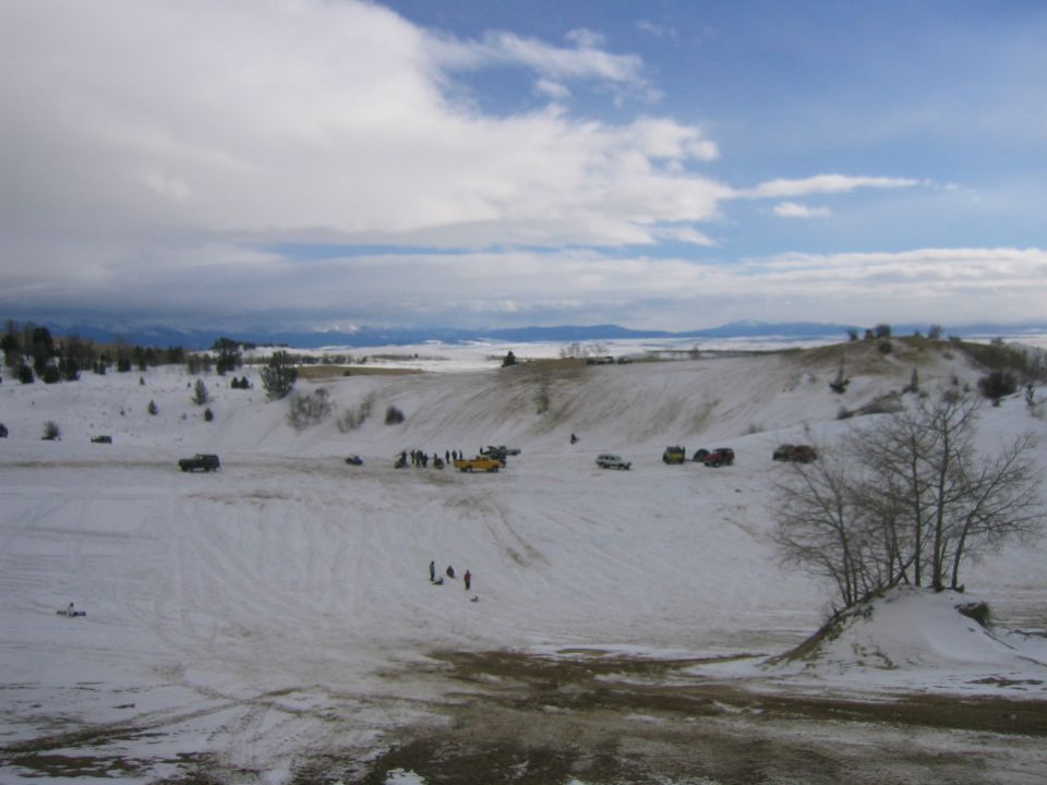 Snowy landscape with several vehicles and groups of people scattered across hills under a cloudy sky