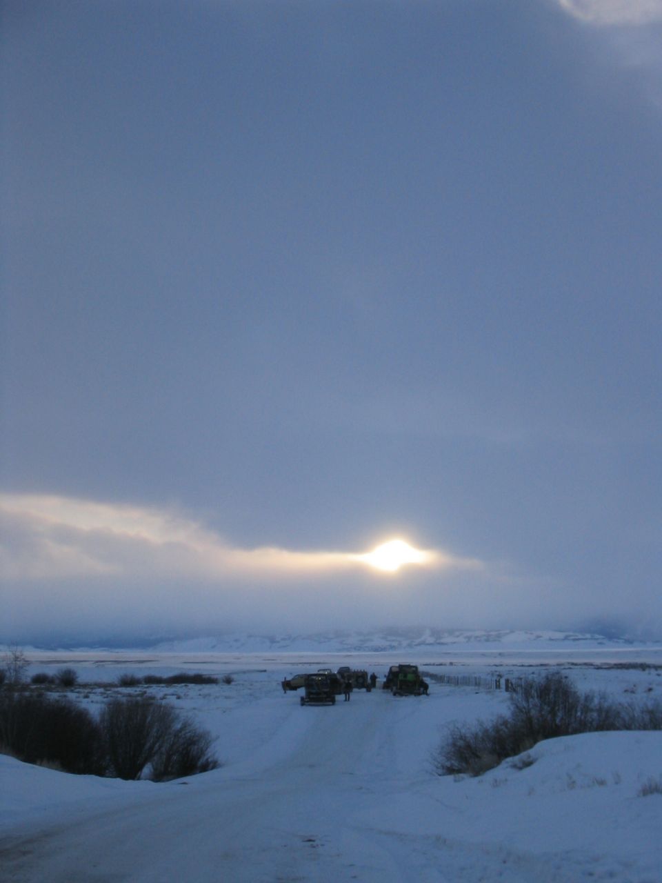 Several vehicles parked in a snowy field with the sun low in the cloudy sky