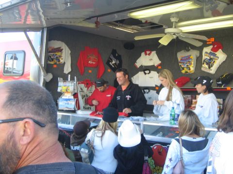 1990 Ford Bronco - Customers crowd a vendor trailer counter lined with hanging T-shirts, hats and jackets, with staff serving behind the glass case.