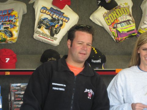 1990 Ford Bronco - man in a black Jim Beam Racing jacket stands behind a vendor counter backed by hanging racing T-shirts and caps.