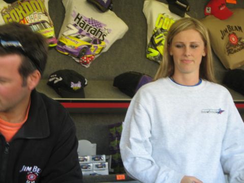 1990 Ford Bronco - blonde woman in a gray sweatshirt and a man in a Jim Beam jacket stand at a merchandise counter with hanging caps and shirts.
