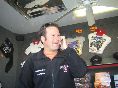 1990 Ford Bronco - man in a black Jim Beam Racing jacket talks on a cellphone inside a merchandise trailer hung with racing shirts and caps.