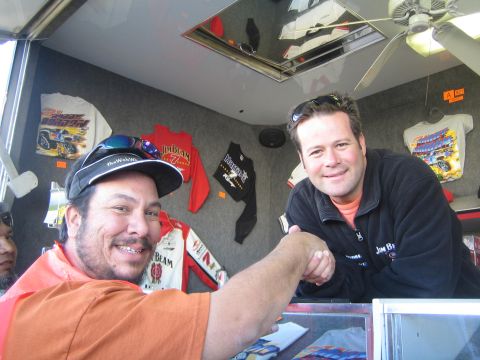 1990 Ford Bronco - Two smiling men shake hands across a glass counter in a racing merchandise trailer hung with shirts and caps.