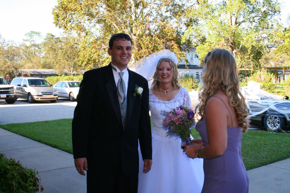 Groom, bride, and bridesmaid stand outside with cars in the background