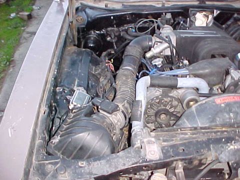 1973 Ford Ranchero - An engine bay view showing a dusty V-configuration engine with a black air cleaner housing, corrugated air intake tubing, radiator hoses, a…
