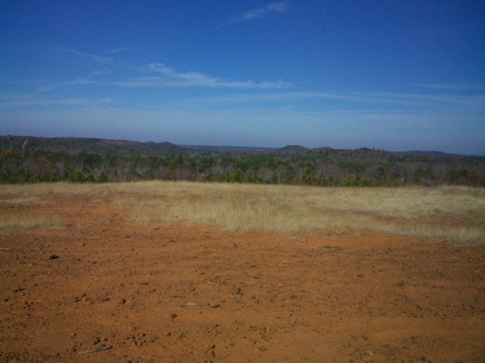 Red dirt clearing and dry grass field overlooking wooded hills under a blue sky