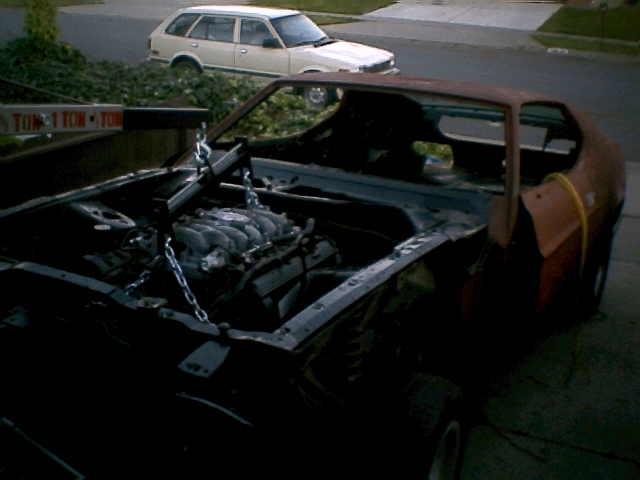 Engine being lifted from rusty car; white station wagon in background