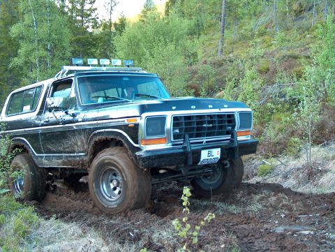1979 Ford Bronco - This Ford Bronco is navigating a muddy path in a forest setting, with trees and foliage all around.