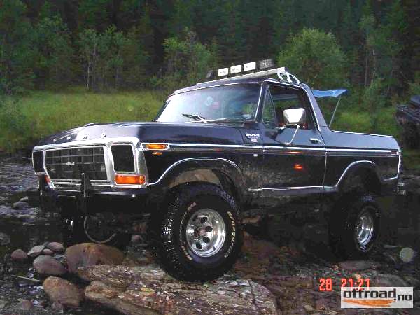 Lifted dark Ford Bronco with roof lights and chrome wheels parked on rocks near a forest stream