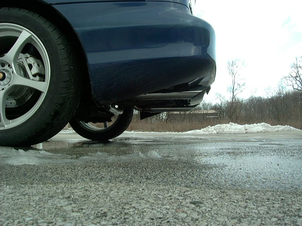 Rear bumper and wheel of a dark blue car, parked on a wet, icy road with snow piles in the background