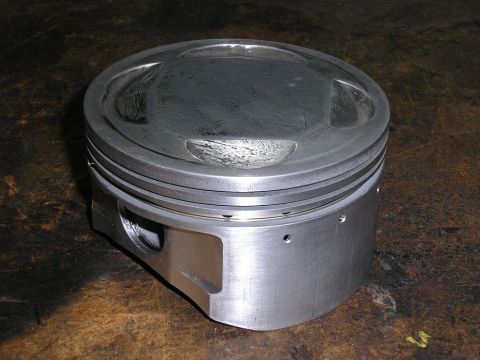 1900 Honda Big Red - This engine piston is shown resting on a textured surface, with visible machining marks and small oil holes.