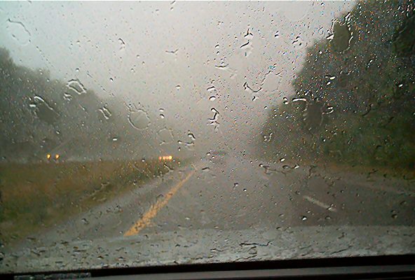 View through a rain-streaked windshield of a two-lane road with yellow dividing lines, trees lining the sides, and distant headlights