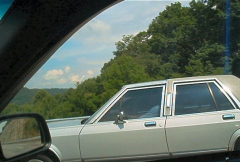 1985 Ford LTD Crown Victoria - A silver 1980s Ford sedan is seen from the passenger side window while driving on a road with a scenic, tree-covered landscape.