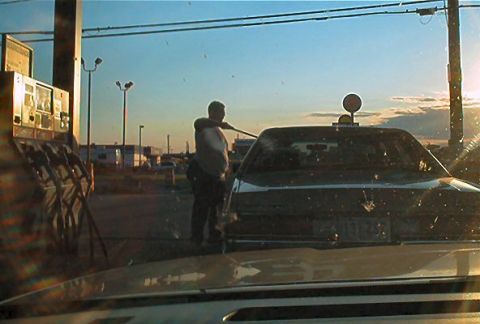 1985 Ford LTD Crown Victoria - A person cleans the rear window of a dark sedan at a gas station during sunset.
