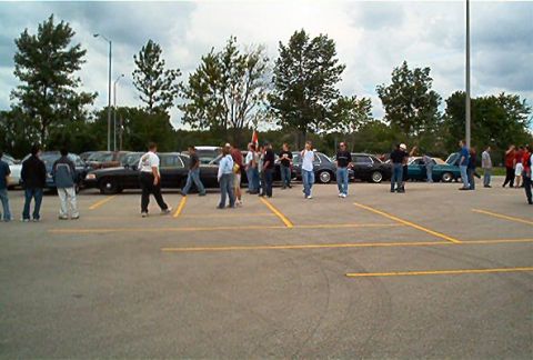 1985 Ford LTD Crown Victoria - A gathering of people and vehicles in a parking lot, with a limousine as a prominent feature.