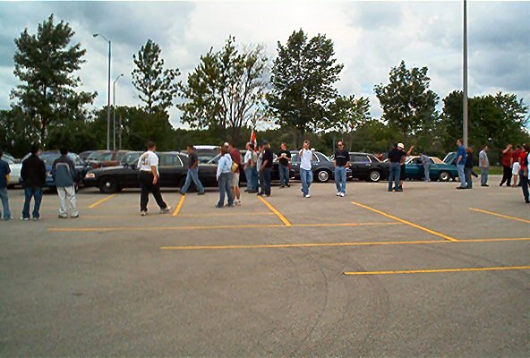 Parking lot scene with people gathered around several cars, including a black limousine and other sedans