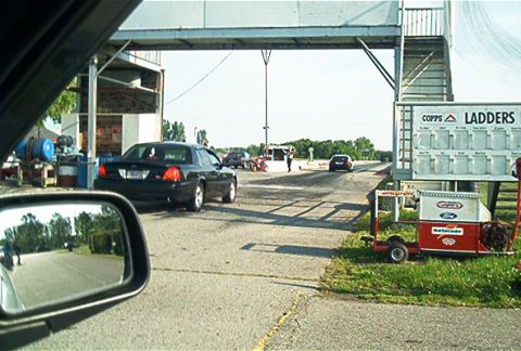 1985 Ford LTD Crown Victoria - A black sedan is seen at a drag strip, with a ladder board displaying race results in the foreground.