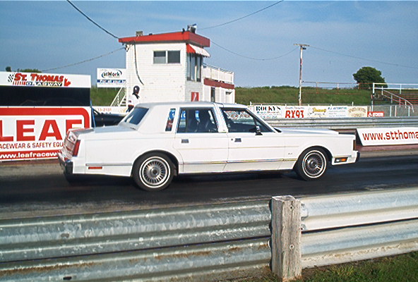 1985 Ford Crown Victoria LTD on a drag strip, side view. Signs for St. Thomas Dragway and Leaf Racewear are visible